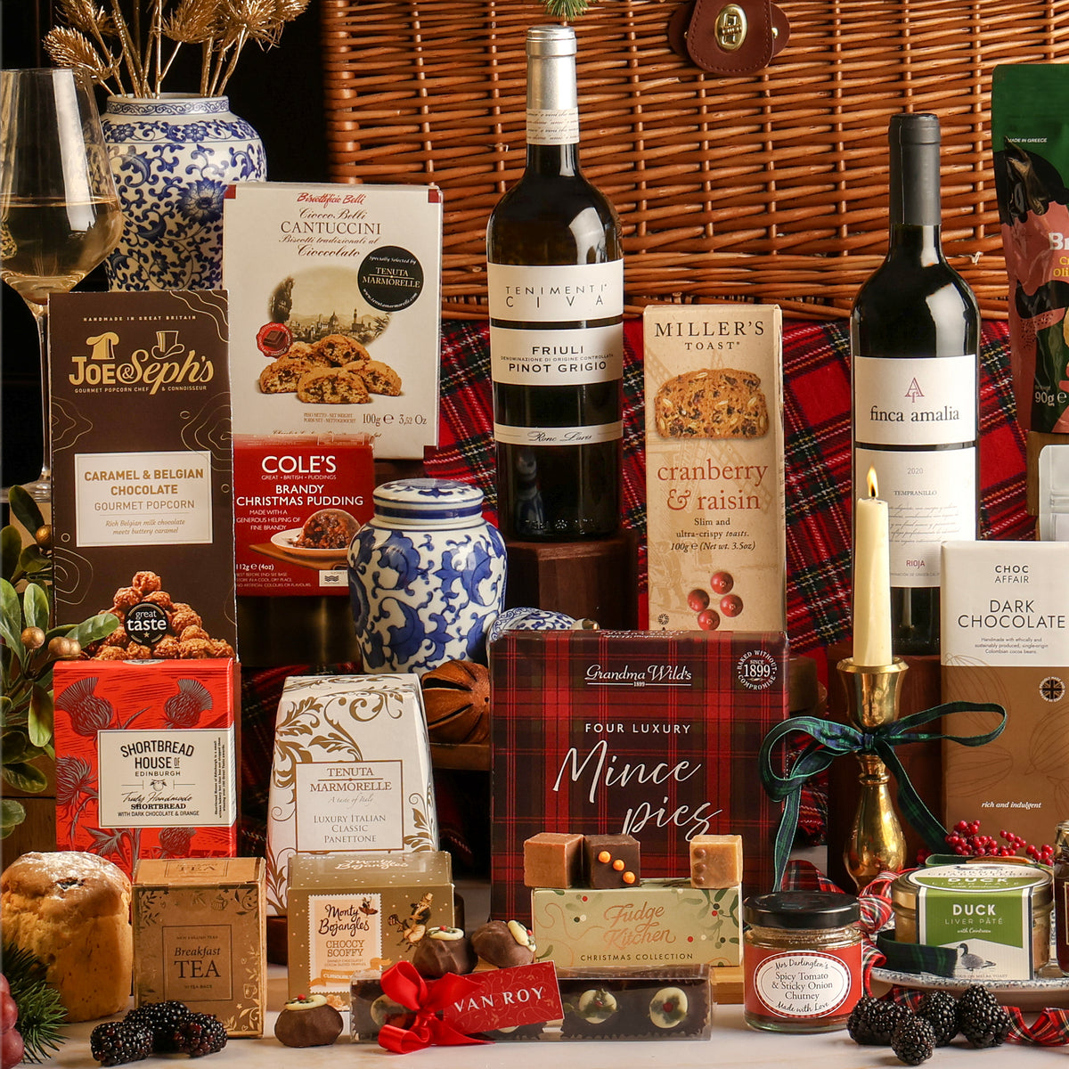 A luxury traditional Christmas hamper gift set arranged in front of a large basket, featuring boxed treats, wine bottles, chocolates, fudge, and small gift items on a white surface.