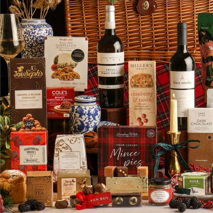 A luxury traditional Christmas hamper gift set arranged in front of a large basket, featuring boxed treats, wine bottles, chocolates, fudge, and small gift items on a white surface.