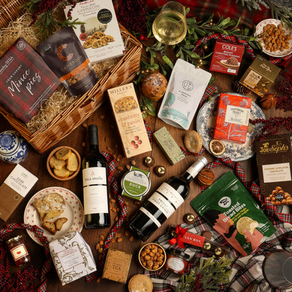 A festive, traditional gift hamper styled in a flat lay on a wooden table, featuring an assortment of gourmet snacks, tea, crackers, and wine. The arrangement includes a basket filled with shredded paper and treats, next to bottles of red and white wine, boxes of biscuits and popcorn, jars of preserves, all decorated with plaid fabrics, pine branches, and red berries.