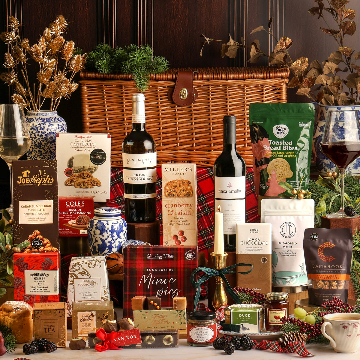 The Traditional Christmas gift hamper featuring a large basket and assorted gourmet treats, including bottles of wine, boxed chocolates, biscuits, and seasonal snacks, styled with dried fruit, red plaid fabric, pine branches, and festive decor.