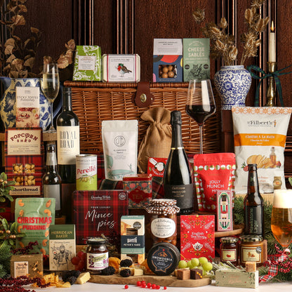 A Luxury Family Sharing Christmas Hamper featuring a wicker basket, assorted gourmet food and drink products, wine, Prosecco, preserves, crackers, and gift boxes, styled on a table with seasonal decorations against a dark wood panelled background.