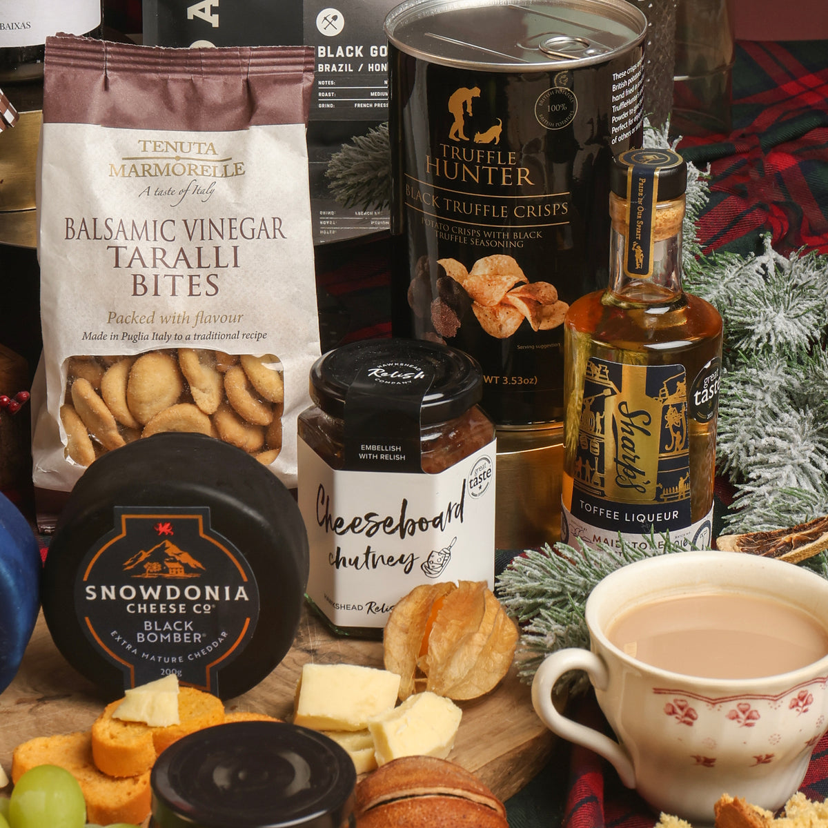 Showcasing a selection of treats from the Magnificent Christmas Hamper, including a bag of Tenuta Marmorelle Balsamic Vinegar Taralli Bites, a can of Truffle Hunter Black Truffle Crisps, a bottle of Toffee Liqueur, a jar of Cheeseboard Chutney, a wheel of Snowdonia Cheese Co. cheese, styled with a cup of tea on a wooden surface surrounded by festive greenery and dried citrus.