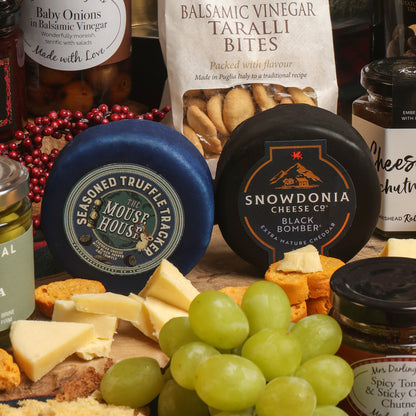 A styled cheese and charcuterie spread with multiple cheeses, crackers, grapes, and jars. Prominent products include a blue-wrapped 'The Mouse House' seasoned truffle cheese, a black-wrapped 'Snowdonia Cheese Co' cheddar cheese, and a bag of Balsamic Vinegar Taralli Bites.