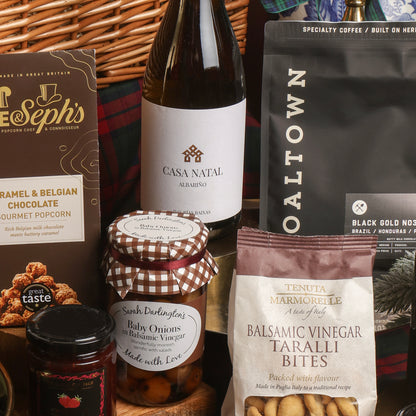 A curated Christmas gift hamper containing multiple packaged food items, including a bottle of Casa Natal wine, a bag of Coaltown Black Gold coffee, Joe and Seph's caramel and Belgian chocolate gourmet popcorn, Tenuta Marmorelle balsamic vinegar taralli bites, Sarah Darlington's baby onions in balsamic vinegar, arranged in front of a wicker basket.
