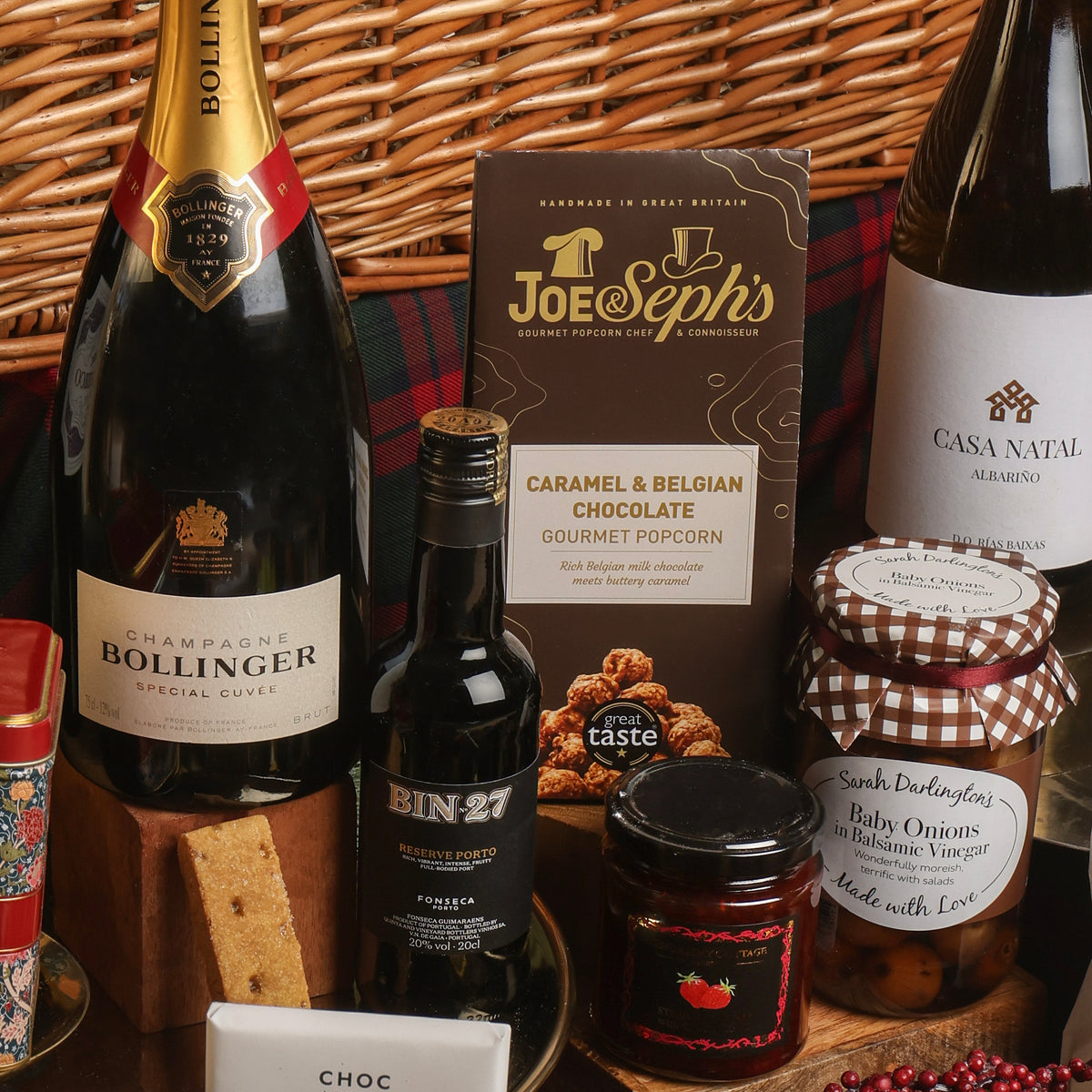 A gourmet gift hamper arrangement featuring a bottle of Bollinger Champagne, a bottle of Bin-27 port, a box of Joe and Seph's gourmet popcorn, a jar of Pickled Baby Onions, and other treats, set against a wicker basket.
