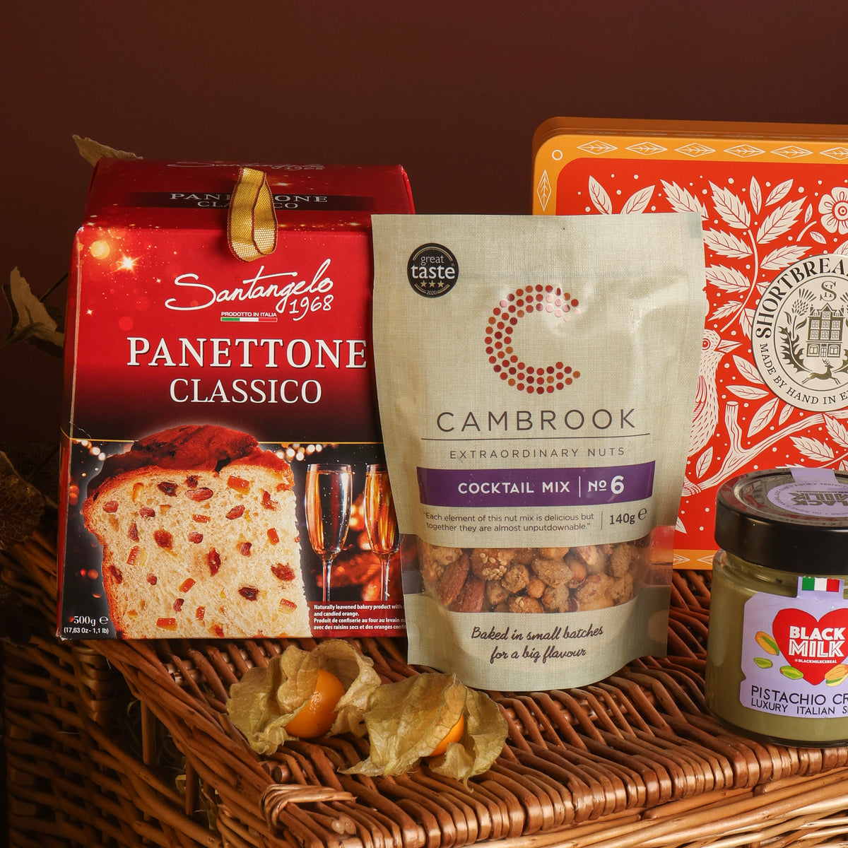 A styled product display featuring a bag of Cambrook Cocktail Mix Nuts in the centre, with a red box of Santangelo Panettone behind, along with a red tin of shortbread and a jar of pistachio spread, all arranged on a wicker basket.