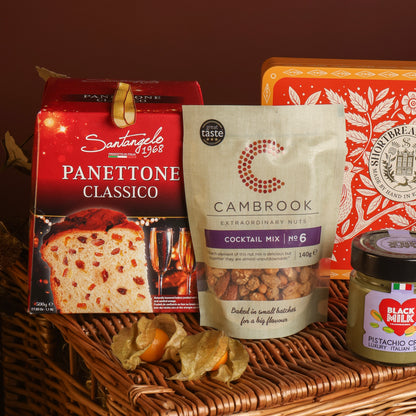 A styled product display featuring a bag of Cambrook Cocktail Mix Nuts in the centre, with a red box of Santangelo Panettone behind, along with a red tin of shortbread and a jar of pistachio spread, all arranged on a wicker basket.