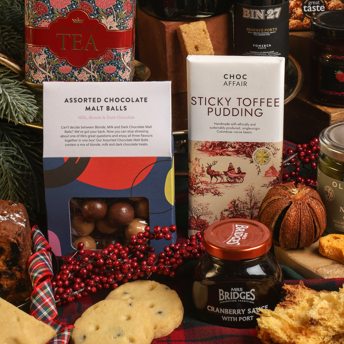 A festive gift assortment of sweet treats, featuring a bar of Choc Affair Sticky Toffee Pudding chocolate, a box of Assorted Chocolate Malt Balls, a jar of cranberry sauce, surrounded by Christmas decorations including red berries and dried fruit on a plaid runner.