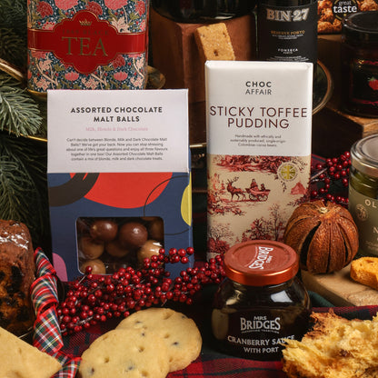 A festive gift assortment of sweet treats, featuring a bar of Choc Affair Sticky Toffee Pudding chocolate, a box of Assorted Chocolate Malt Balls, a jar of cranberry sauce, surrounded by Christmas decorations including red berries and dried fruit on a plaid runner.