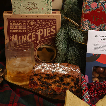 A Christmas-themed selection of food and drink from The Magnificent Christmas Hamper, featuring a box of Fudge Kitchen fudge, a tin of tea, mince pies, a box of assorted chocolate malt balls, a glass with festive drink and ice, a fruit cake dusted with powdered sugar, and festive decorations including pine branches and plaid fabric.