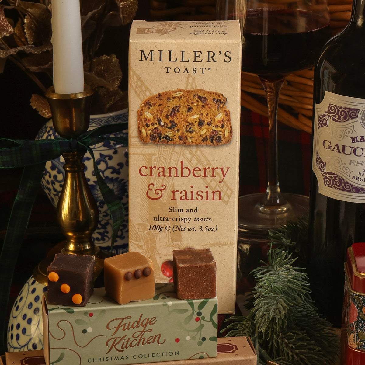 Miller's Toast cranberry and raisin toast box displayed on a tabletop with fudge pieces, wine, and Christmas decorations including a candle, wine glass, and pine branches, in a styled indoor setting.