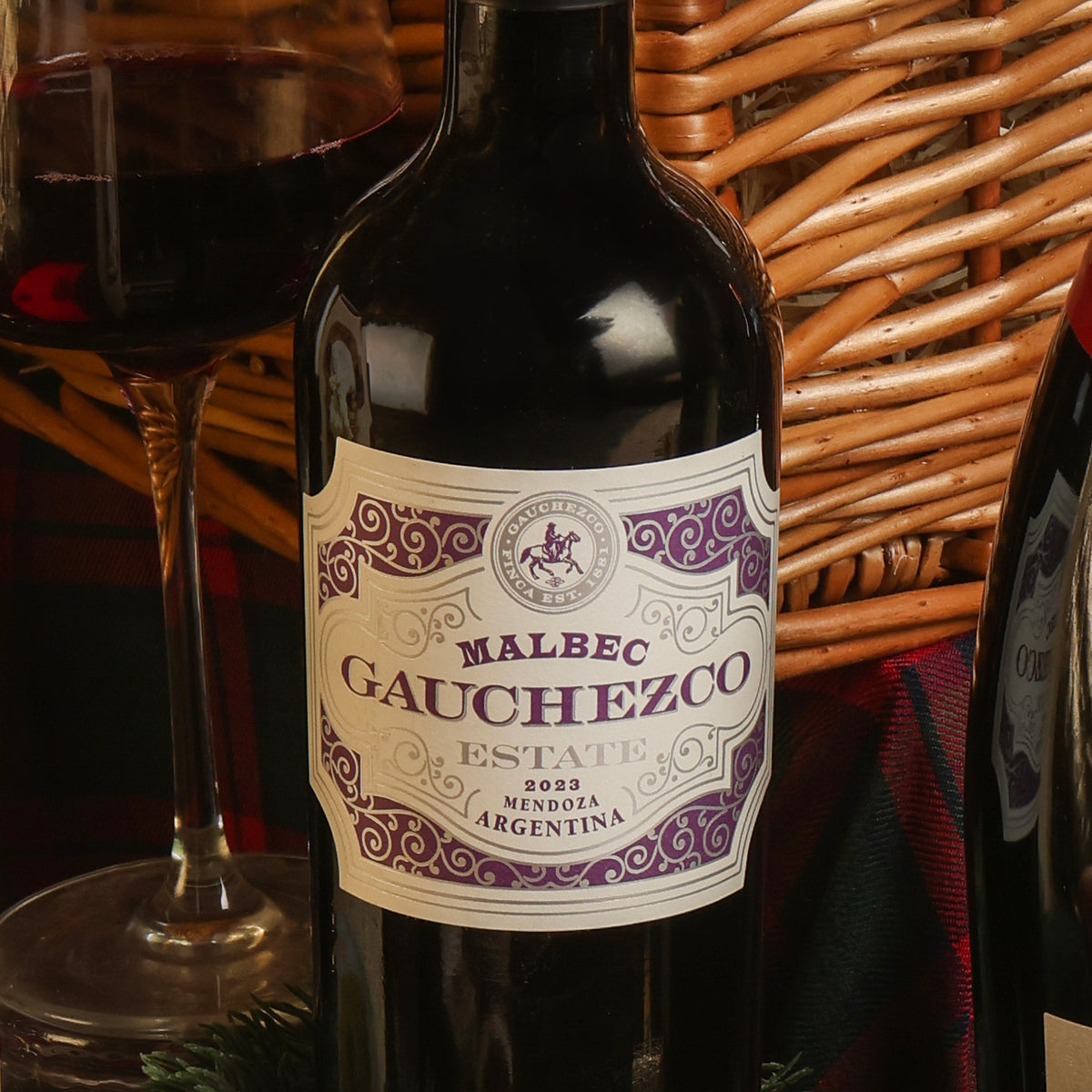 A bottle of Gauchezco Estate Malbec red wine with a cream and purple label, set against a wicker basket with a glass of red wine partially visible on the left.