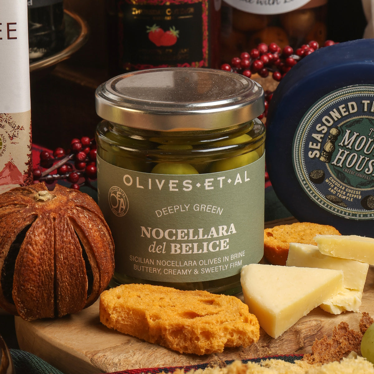 A jar of Olives Et Al Deeply Green Sicilian olives is styled on a wooden board with crackers, cheese, and a round blue cheese wheel in a rustic, warm setting.