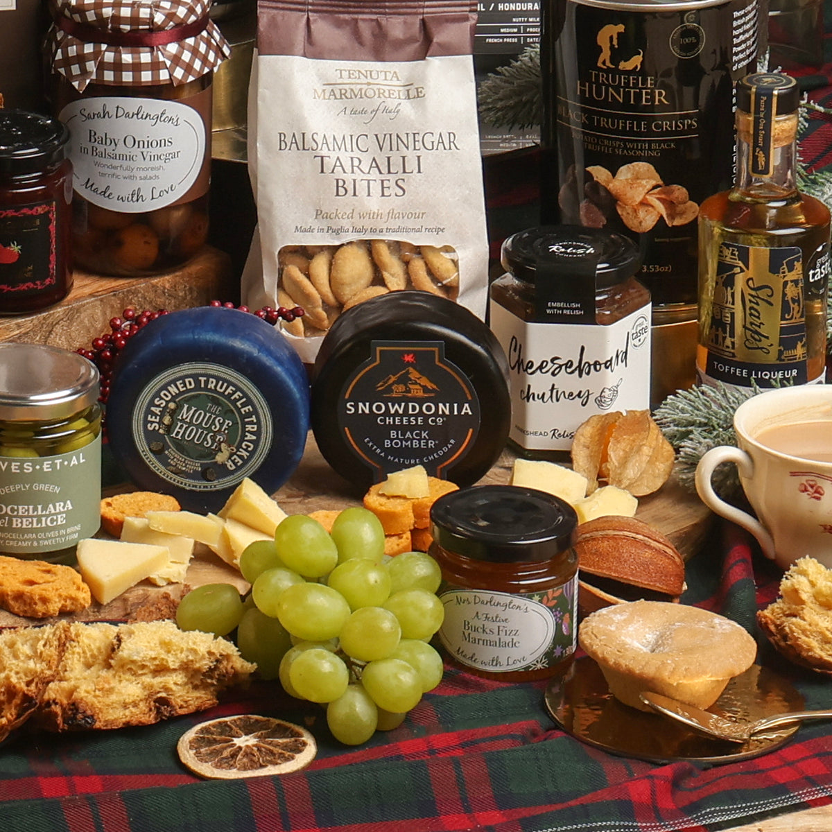 A styled Christmas cheese and charcuterie spread on a plaid tablecloth, featuring an assortment of cheeses, crackers, green grapes, preserves, and gourmet snacks from brands like Tenuta Marmorelle, Snowdonia Cheese Co, and The Mouse House.