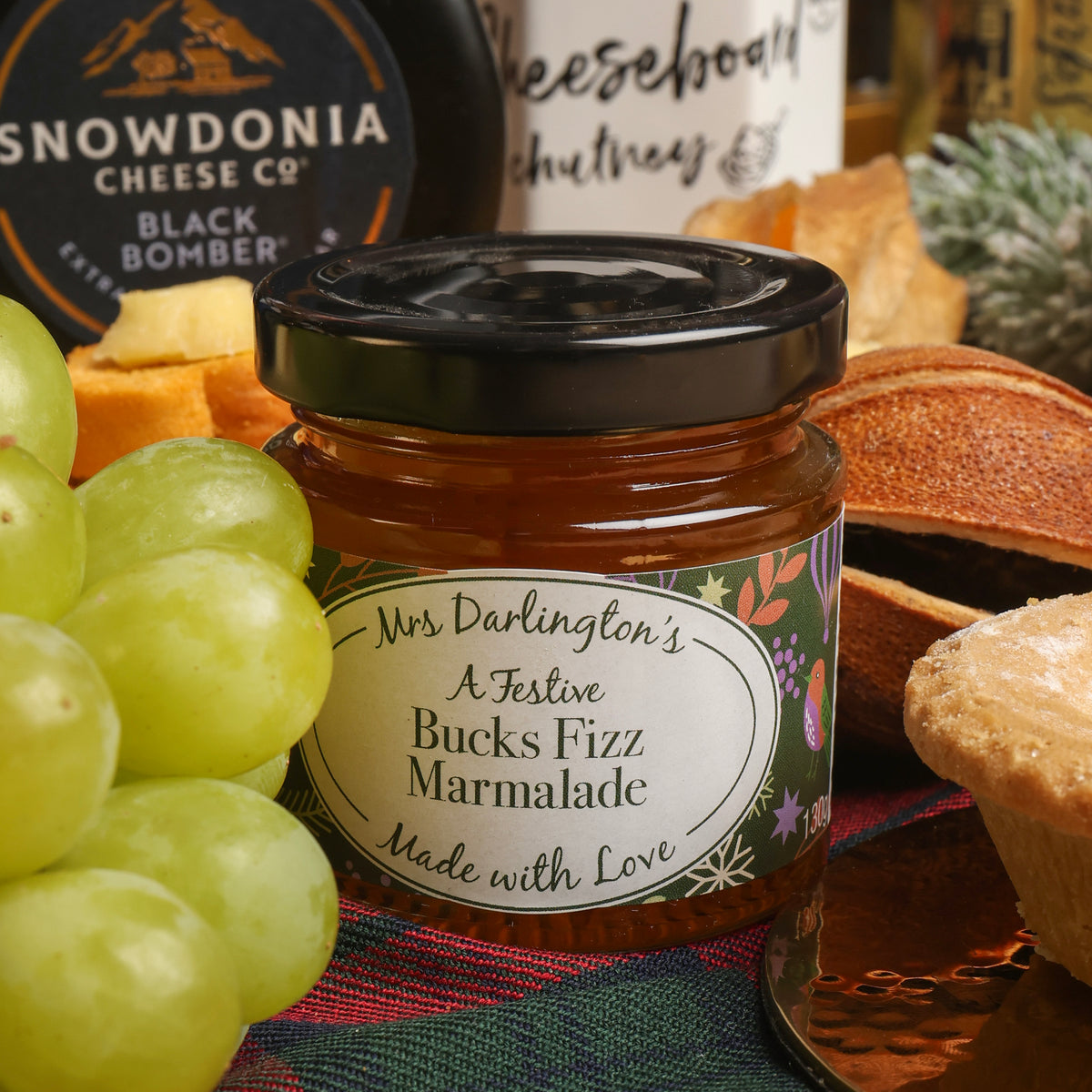 A glass jar of Mrs Darlington's Bucks Fizz Marmalade with a black lid, surrounded by festive food props including green grapes, cheese board chutney, and a mince pie, on a patterned plaid cloth.