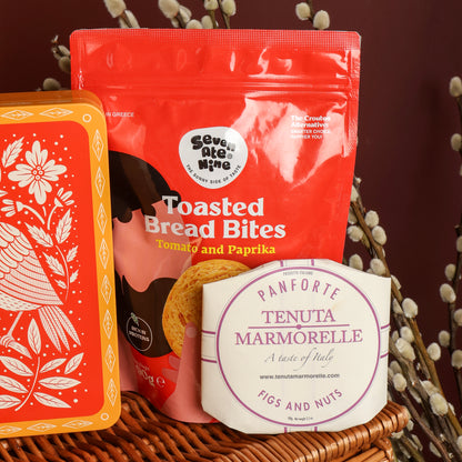 Close-up of items from the Magnificent Christmas Hamper, sat on top of the wicker hamper, featuring a red stand-up pouch of Toasted Bread Bites, a round white package of Tenuta Marmorelle Panforte Figs and Nuts, and a decorative orange tin with white floral and bird illustrations, styled with dried branches against a dark brown background.