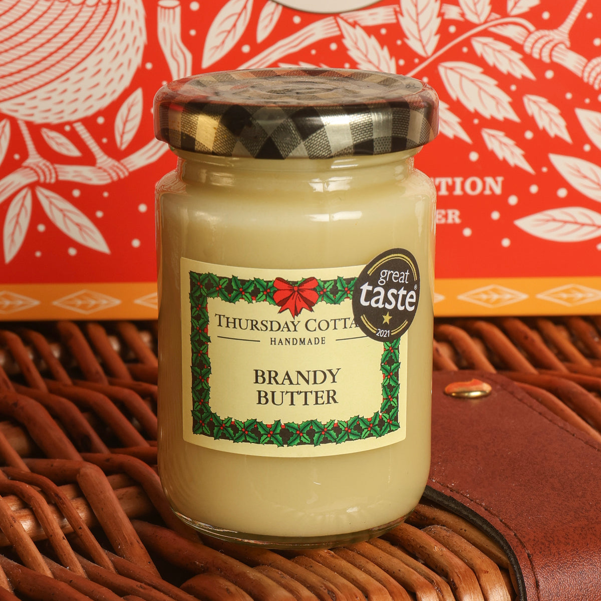 A glass jar of Thursday Cottage Handmade Brandy Butter with a black-and-white plaid lid, placed on a brown wicker basket with a red patterned tin in the background.