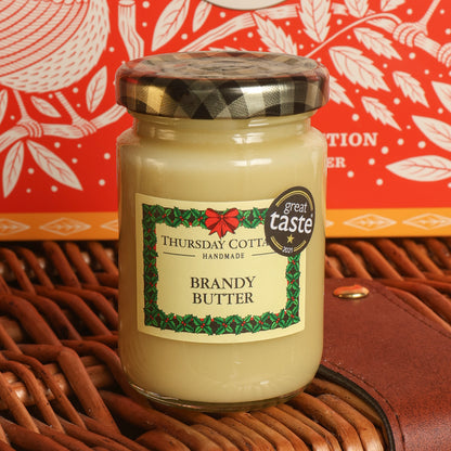 A glass jar of Thursday Cottage Handmade Brandy Butter with a black-and-white plaid lid, placed on a brown wicker basket with a red patterned tin in the background.