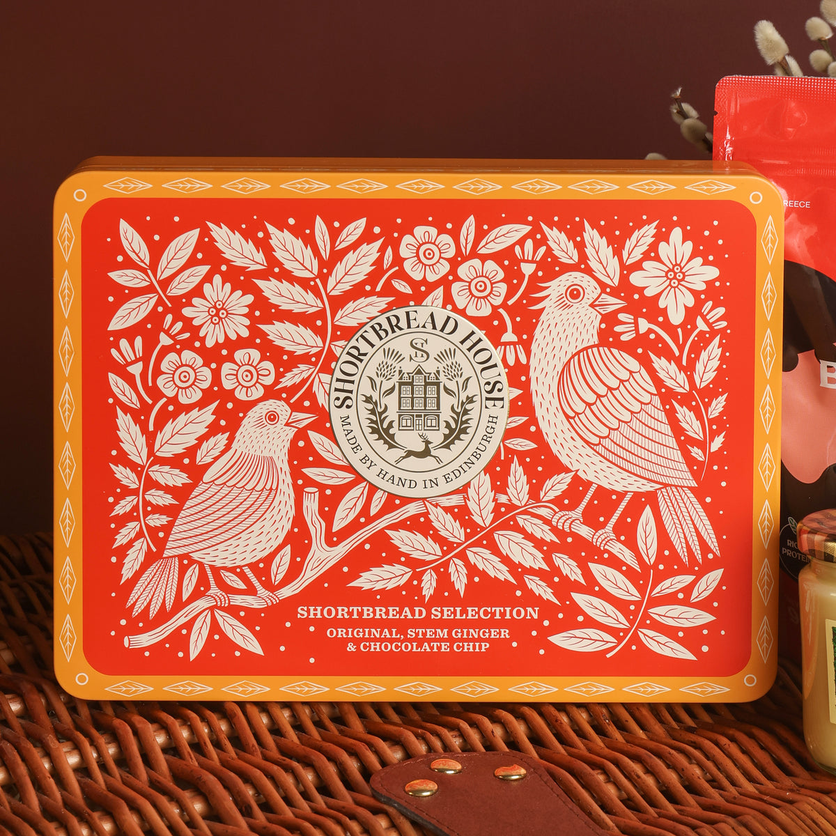 A rectangular red and orange decorative tin box with white floral and bird illustrations, labelled 'Shortbread Selection' by Shortbread House, placed on a woven wicker basket.