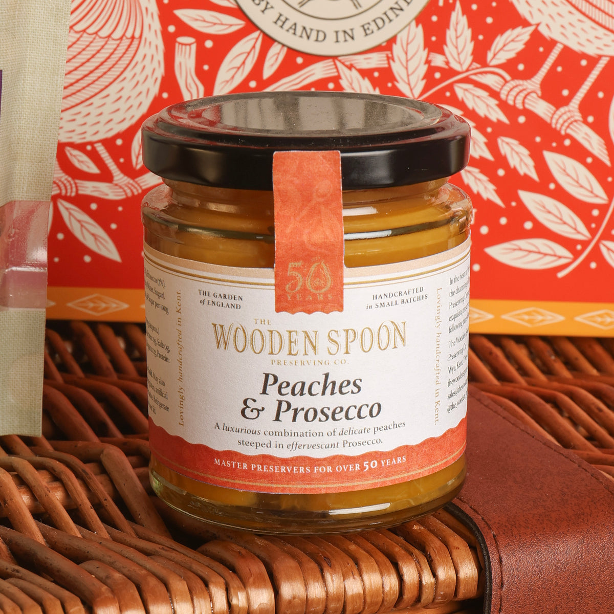 A glass jar of The Wooden Spoon Preserving Co. Peaches & Prosecco preserve, featuring a black lid and a terracotta-coloured paper seal, displayed on a brown wicker basket with a red patterned tin in the background.
