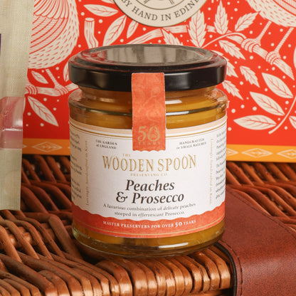 A glass jar of The Wooden Spoon Preserving Co. Peaches & Prosecco preserve, featuring a black lid and a terracotta-coloured paper seal, displayed on a brown wicker basket with a red patterned tin in the background.