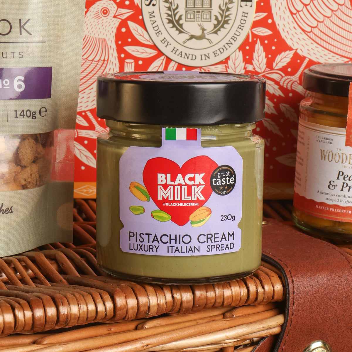A jar of Black Milk's Pistachio Cream Luxury Italian Spread, featuring a black lid and a white label with a red heart logo, displayed on a brown wicker basket with other gourmet items in the background.