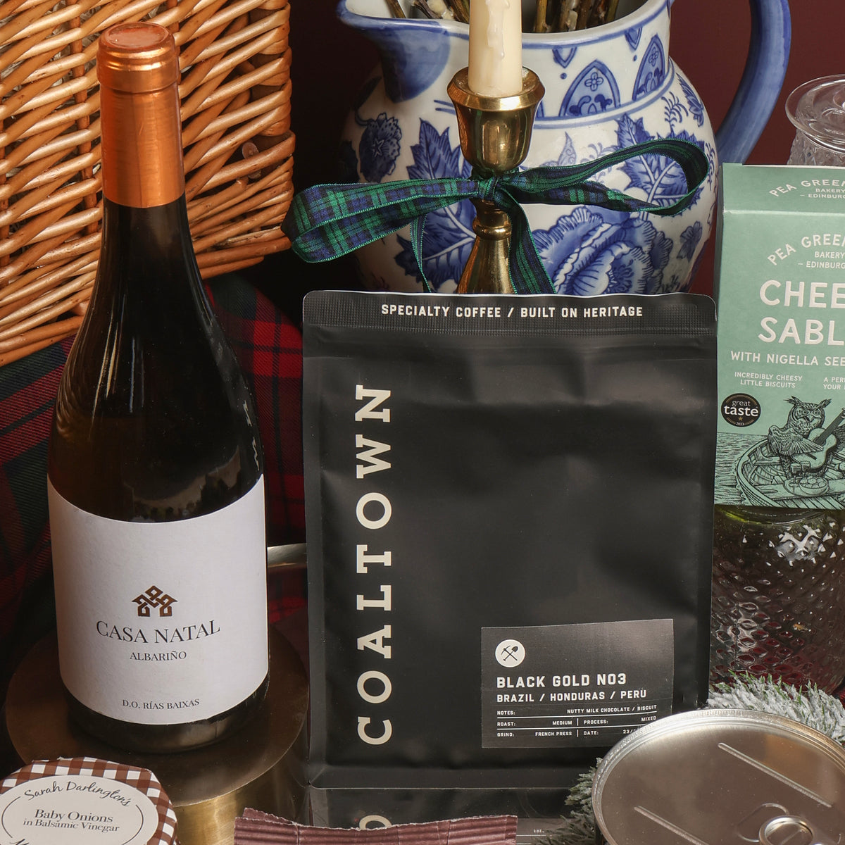 A gift basket arrangement featuring a bottle of Casa Natal white wine, a black bag of Coaltown Black Gold No. 3 specialty coffee, and a green box of Cheese Sablés, styled with a wicker basket, blue and white ceramic jar, and candle.