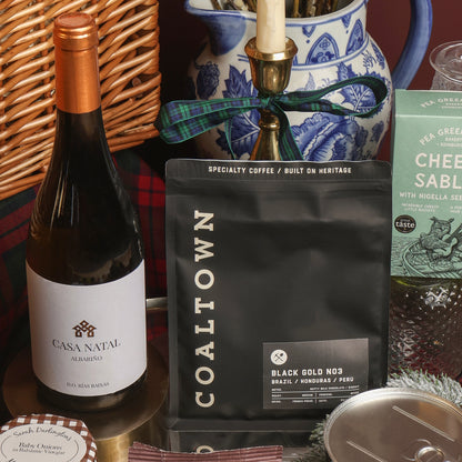 A gift basket arrangement featuring a bottle of Casa Natal white wine, a black bag of Coaltown Black Gold No. 3 specialty coffee, and a green box of Cheese Sablés, styled with a wicker basket, blue and white ceramic jar, and candle.