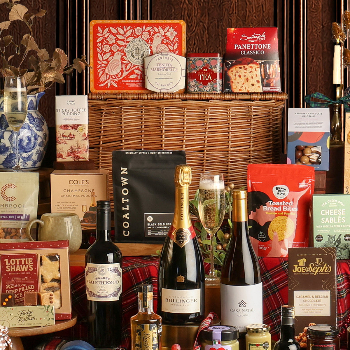 The Magnificent Christmas Hamper gift set with a wicker basket, featuring bottles of wine, Bollinger Champagne, tea, cookies, Cole's Christmas pudding, Lottie Shaw's mince pies, and other packaged snacks on a red plaid fabric.