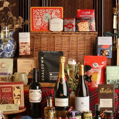 The Magnificent Christmas Hamper gift set with a wicker basket, featuring bottles of wine, Bollinger Champagne, tea, cookies, Cole's Christmas pudding, Lottie Shaw's mince pies, and other packaged snacks on a red plaid fabric.