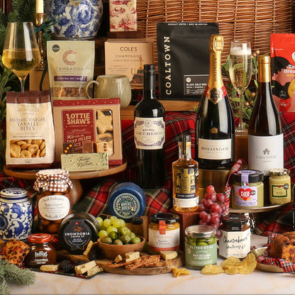 Close-up of multiple items from The Magnificent Christmas Hamper with a charcuterie spread arranged on a table with a plaid runner, in front of a wicker basket. Visible items include bottles of wine and Champagne, packaged snacks, jars of preserves, crackers, cheese, grapes, with a black Coaltown coffee bag and other gift packages in the background.