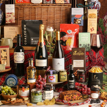 A festive Christmas hamper filled with gourmet food and drink items, including bottles of wine and Champagne, assorted cheeses, crackers, preserves, and chocolates, arranged on a table with red plaid fabric, dried fruits and Christmas decorations.