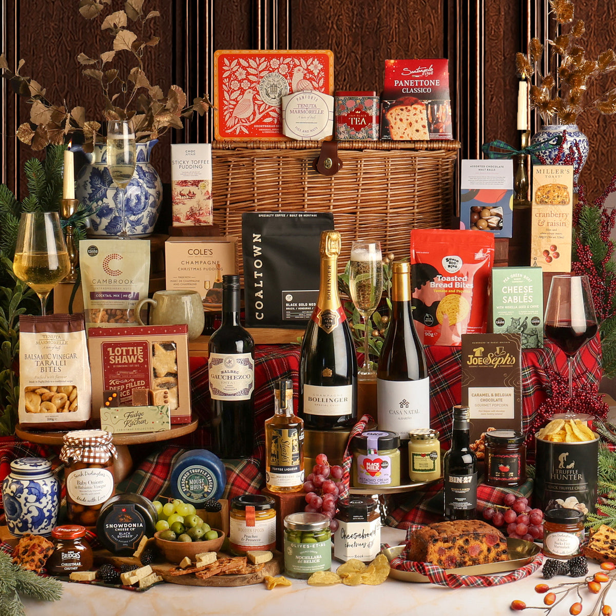 Showcasing all of the food and drink items from The Magnificent Christmas Hamper, surrounding a large wicker basket, featuring assorted gourmet treats, crackers, and sweets, alongside several bottles of wine and Champagne, jars of preserves and spreads, and a Christmas pudding, styled with grapes, cheese wedges, and red tartan fabric against a dark wood panel background.