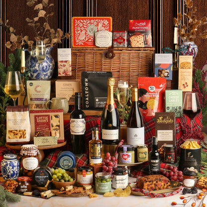 Showcasing all of the food and drink items from The Magnificent Christmas Hamper, surrounding a large wicker basket, featuring assorted gourmet treats, crackers, and sweets, alongside several bottles of wine and Champagne, jars of preserves and spreads, and a Christmas pudding, styled with grapes, cheese wedges, and red tartan fabric against a dark wood panel background.
