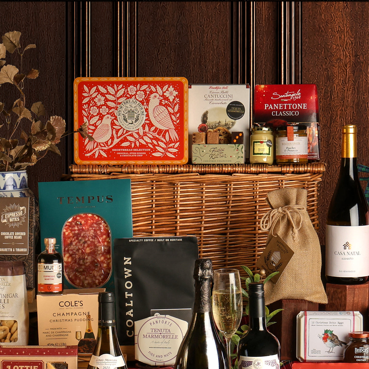Close-up of a few food and drink items included with the large Ultimate Christmas Hamper, featuring a large wicker hamper gift basket and assorted gourmet treats, biscuits, preserves, chocolates, charcuterie, wine bottles, and panettone, arranged against a dark wood-panelled background.