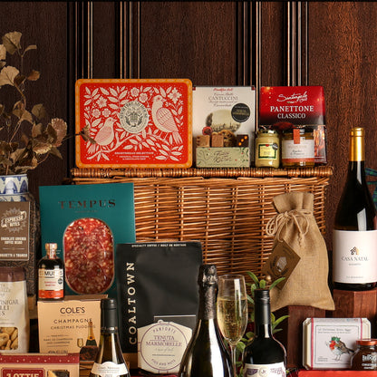 Close-up of a few food and drink items included with the large Ultimate Christmas Hamper, featuring a large wicker hamper gift basket and assorted gourmet treats, biscuits, preserves, chocolates, charcuterie, wine bottles, and panettone, arranged against a dark wood-panelled background.