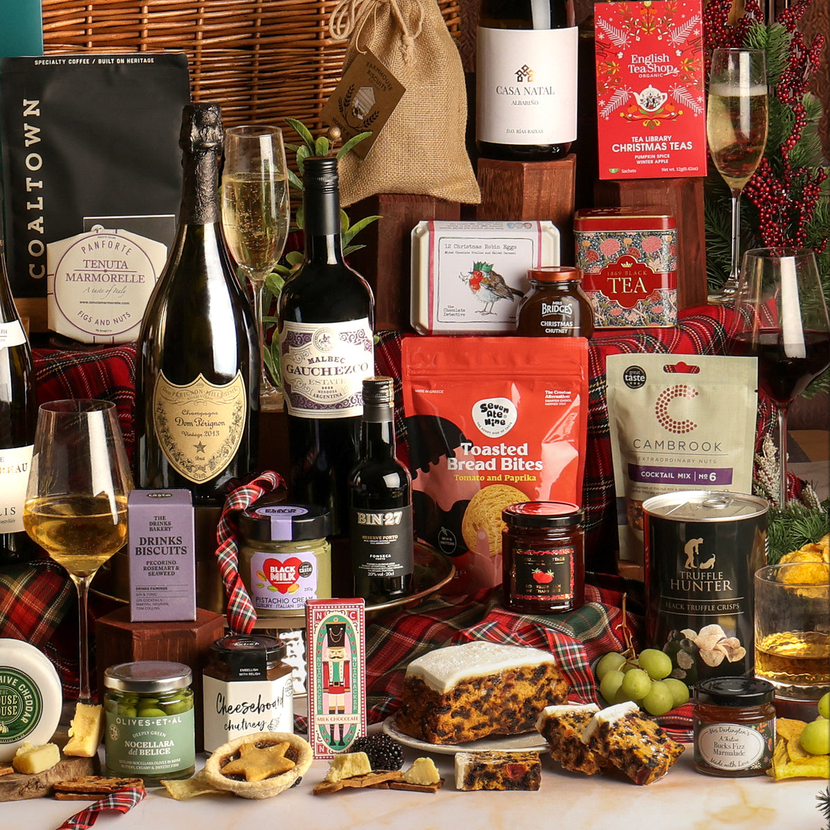 Close-up of multiple items from The Ultimate Christmas Hamper, displayed as a festive gift hamper arrangement featuring an assortment of food and beverage products including wine, tea, cake, cheeses, preserves, and a loaf of cake, styled with a glass filled with Prosecco, plaid fabric, red berries, and festive decorations.