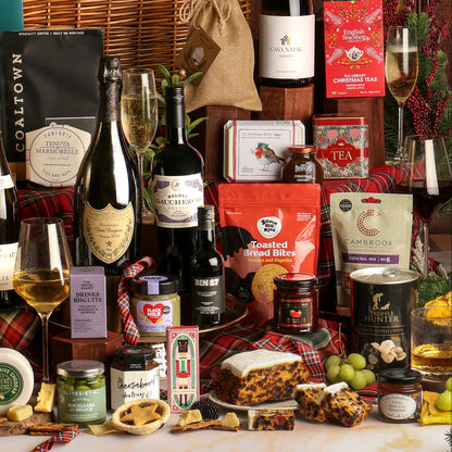 Close-up of multiple items from The Ultimate Christmas Hamper, displayed as a festive gift hamper arrangement featuring an assortment of food and beverage products including wine, tea, cake, cheeses, preserves, and a loaf of cake, styled with a glass filled with Prosecco, plaid fabric, red berries, and festive decorations.