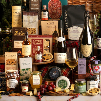 A luxury gourmet gift hamper filled with an assortment of premium foods and beverages, including bottles of wine, Champagne, cheeses, crackers, preserves, and chocolates, arranged on a table with a plaid tartan runner and Christmas decorations.