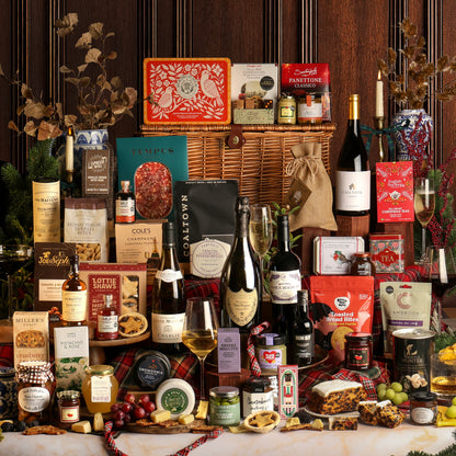 The Ultimate Christmas Hamper with a large assortment of gourmet foods and drinks designed to share, featuring cheeses, crackers, wine bottles, mince pies, chocolates, and other packaged snacks arranged around a large wicker basket on a table. The indoor setting includes Christmas decor in the background.