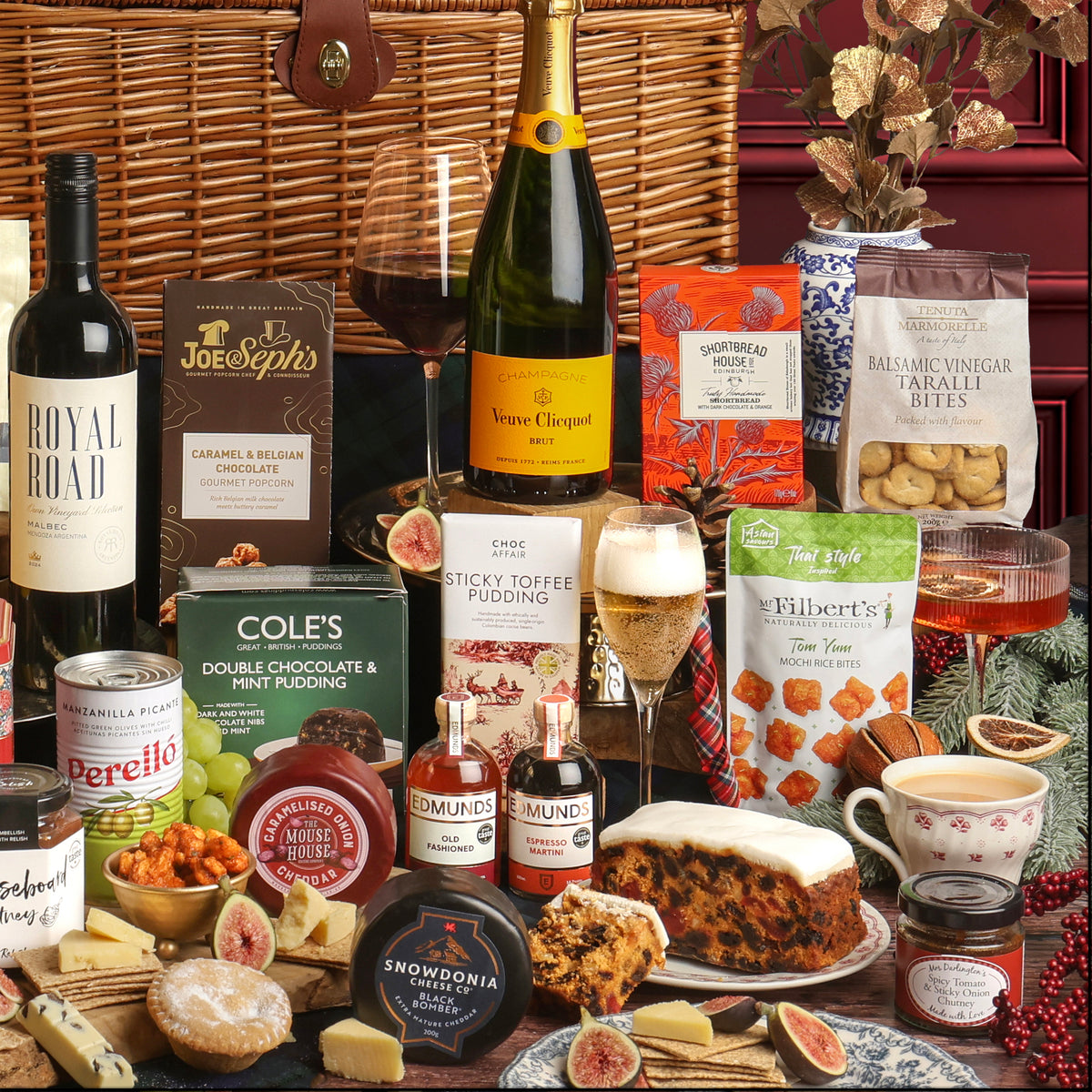 A luxury Christmas Celebration Hamper arranged on a wooden table, featuring a bottle of wine, Champagne, several packaged snacks and sweets, and cheeses, styled with figs and berries in a festive setting.
