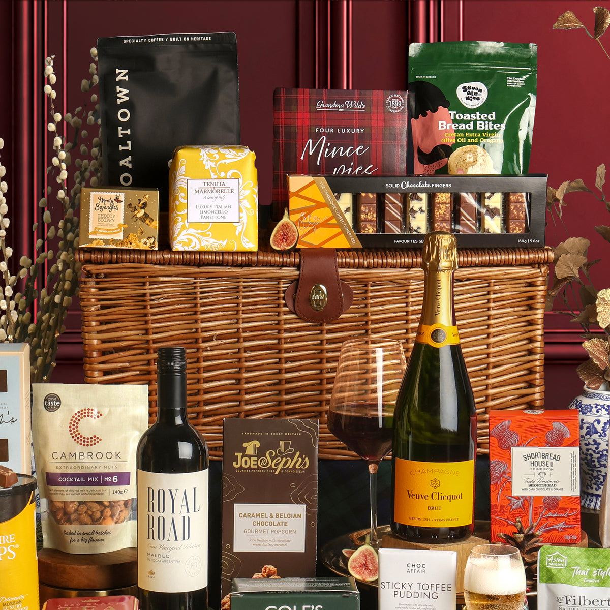 The Christmas Celebration Hamper styled with a dark red wooden panel background, featuring drinks and snacks surrounding the hamper, including red wine, coffee, nuts, chocolate fingers and shortbread.