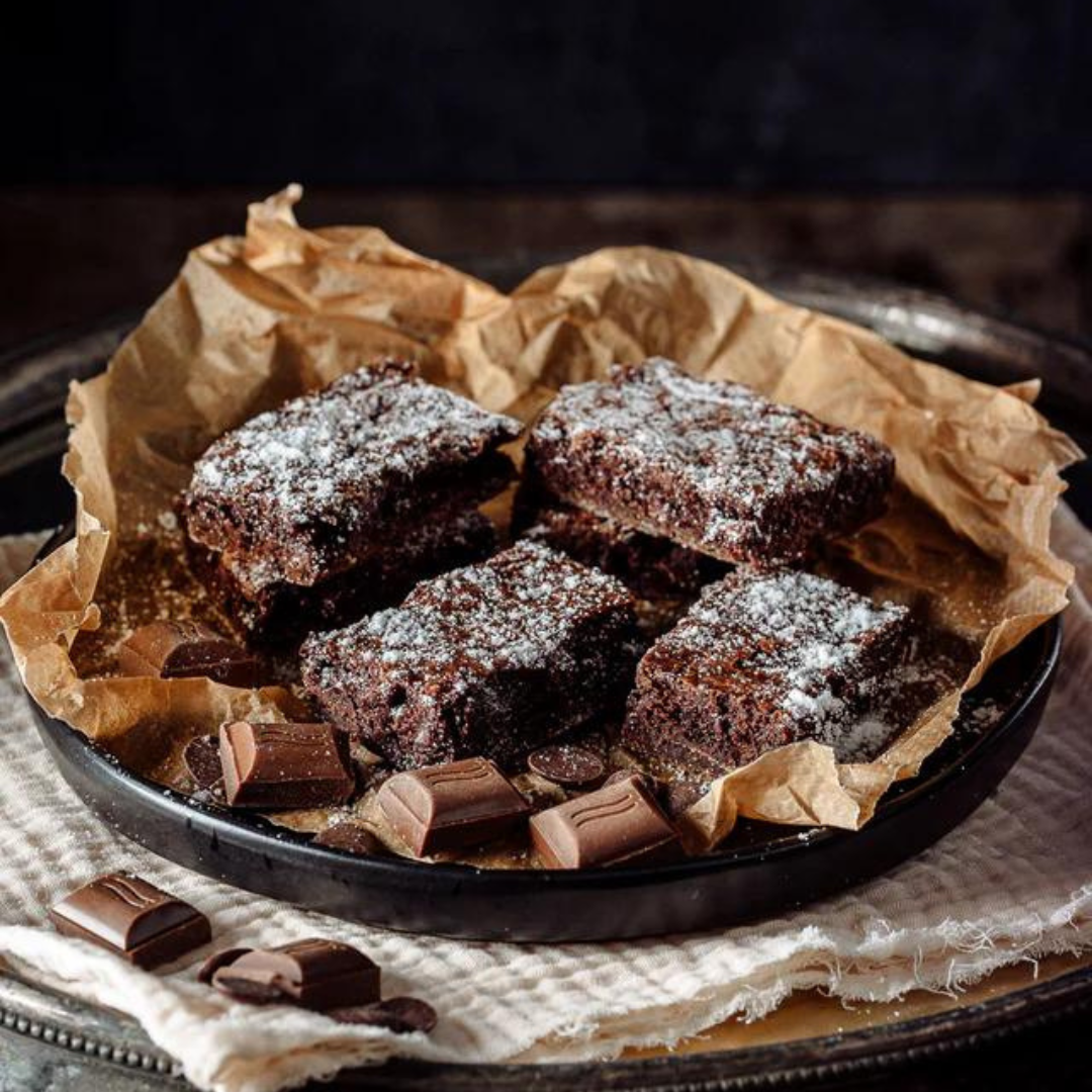 Four Belgian chocolate brownies handmade by Wilfreds, dusted with powdered sugar, arranged on a black plate lined with brown parchment paper, with chocolate pieces scattered around on a cloth-covered tray.