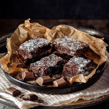 Four Belgian chocolate brownies handmade by Wilfreds, dusted with powdered sugar, arranged on a black plate lined with brown parchment paper, with chocolate pieces scattered around on a cloth-covered tray.