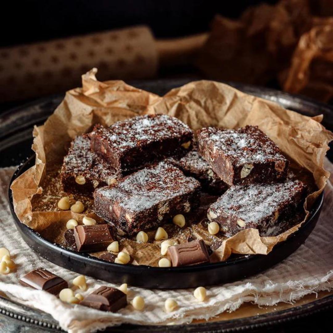Several pieces of chocolate chip brownie dusted with powdered sugar, arranged on parchment paper inside a black dish with chocolate pieces and white chocolate chips around them.