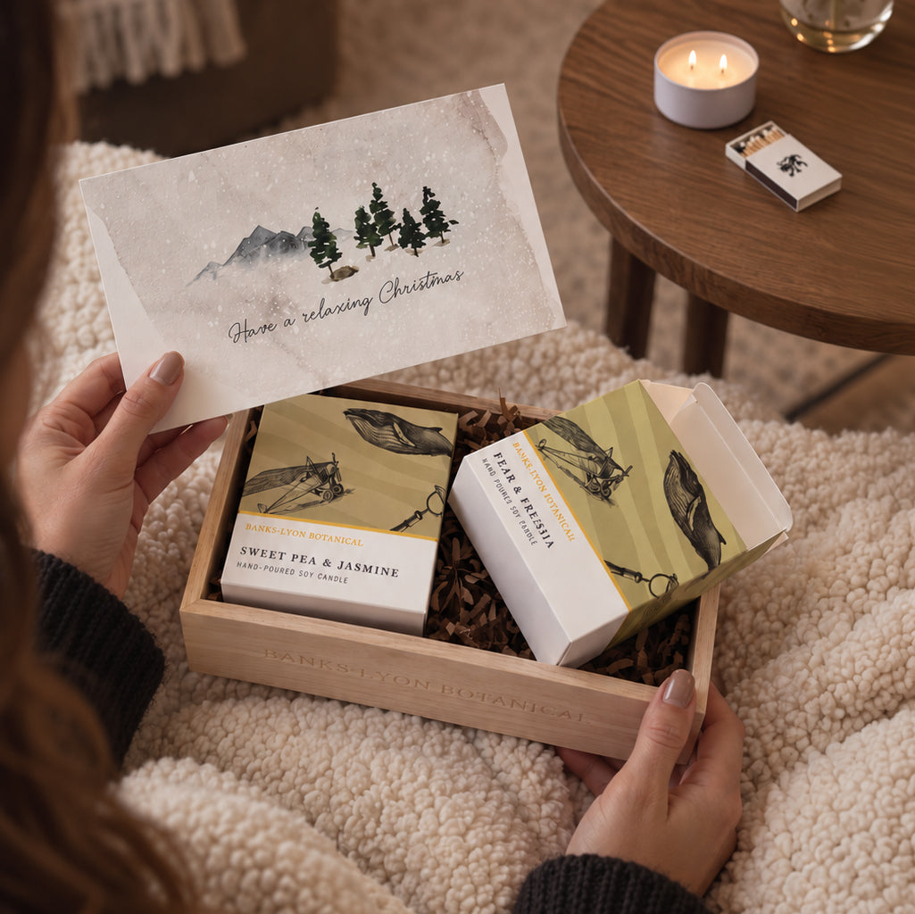 A wooden gift box opened to reveal two soy candles in a Christmas candle gift set, with a person holding the gift.