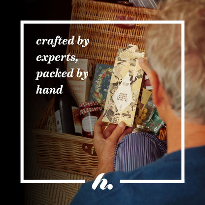 An informative USP image showing a person viewed from behind reaching into a wicker basket hamper filled with packaged food items, holding a cream-coloured box of salted caramel truffles. The image has a white border with the text 'crafted by experts, packed by hand' and a stylised logo at the bottom.