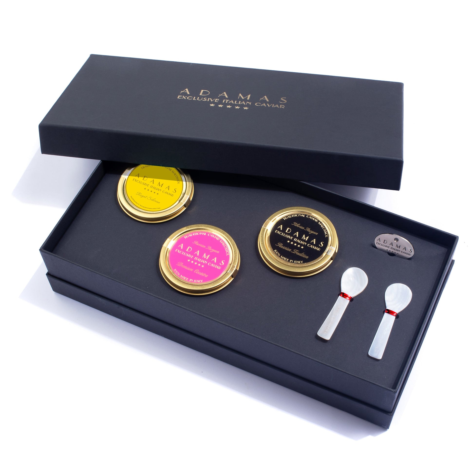 An open Adamas gift box containing a selection of three caviar varieties (yellow, pink, and black labels), a small tin opener, and two mother of pearl spoons, photographed on a white background.
