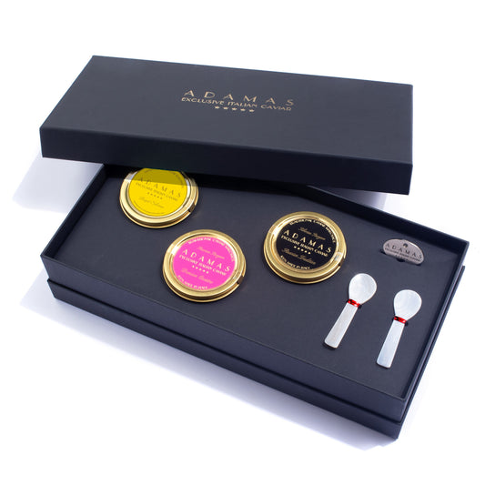 An open Adamas gift box containing a selection of three caviar varieties (yellow, pink, and black labels), a small tin opener, and two mother of pearl spoons, photographed on a white background.