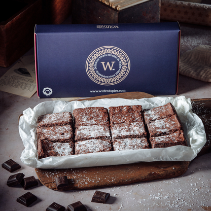 A box of chocolate brownies by Wilfreds sits behind a tray of cut brownies dusted with powdered sugar, arranged on parchment paper over a wooden board with scattered chocolate pieces.